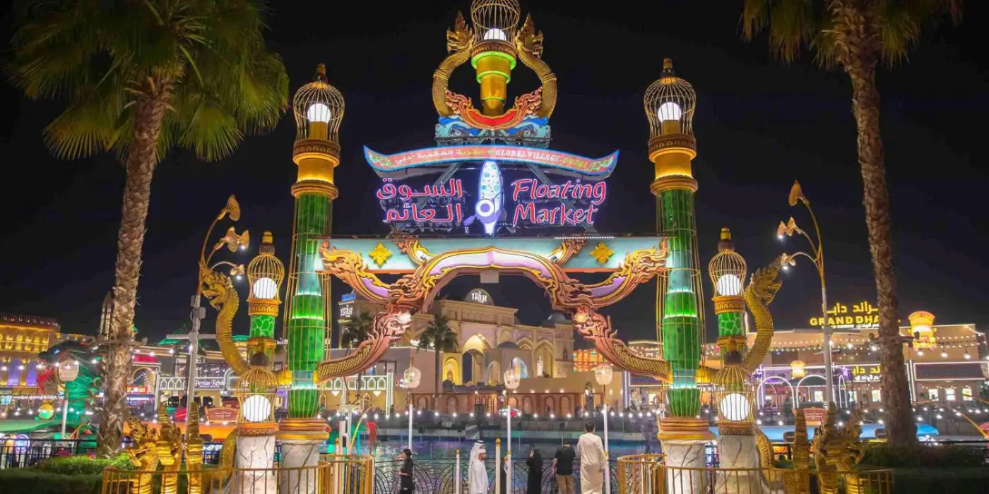 A brightly lit entrance to the Floating Market at Global Village Dubai, with a large archway adorned with colorful lanterns and palm trees on either side.