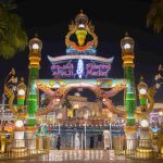 A brightly lit entrance to the Floating Market at Global Village Dubai, with a large archway adorned with colorful lanterns and palm trees on either side.