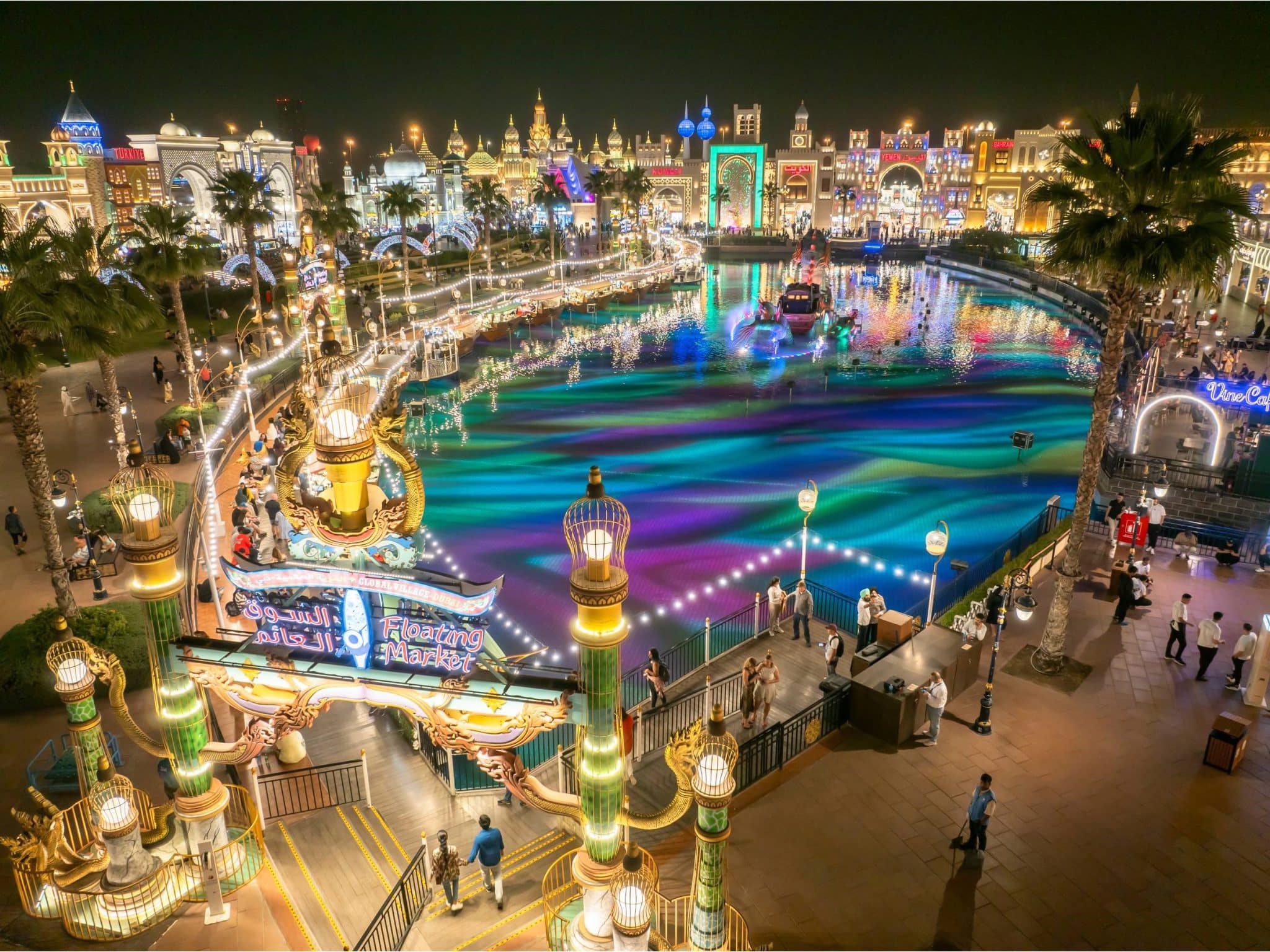 A vibrant aerial view of Global Village Dubai at night. The canal is illuminated with colorful lights, and there are numerous pavilions and attractions visible in the background.