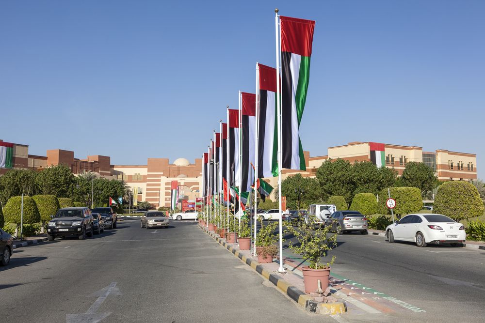 Street view of Al Jurf, Ajman, showcasing a well-maintained road lined with UAE flags, lush greenery, parked cars, and modern buildings in the background