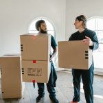 Two professional movers in uniform carrying cardboard boxes in a bright, spacious room with arched windows, symbolizing efficient moving and packing services in Ajman