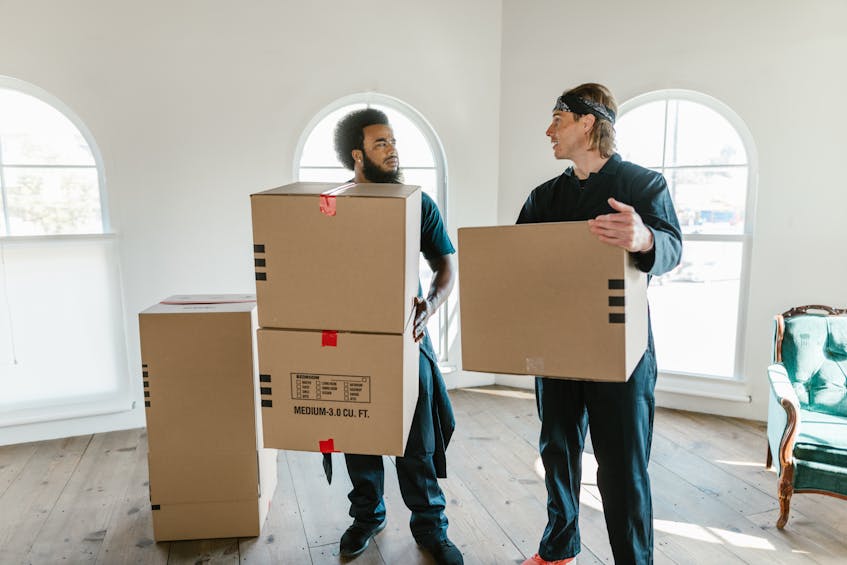 Two professional movers in uniform carrying cardboard boxes in a bright, spacious room with arched windows, symbolizing efficient moving and packing services in Ajman