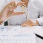 A real estate agent holding a small wooden house with a key, symbolizing the concept of an escrow account in UAE real estate, with financial charts and a laptop in the background