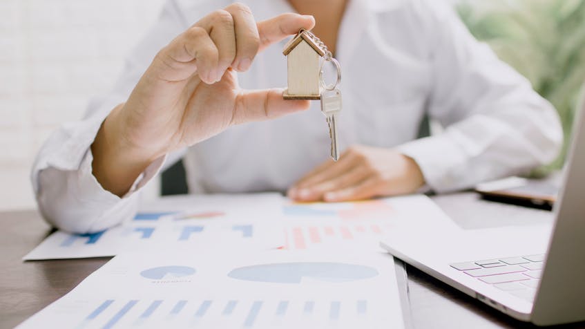 A real estate agent holding a small wooden house with a key, symbolizing the concept of an escrow account in UAE real estate, with financial charts and a laptop in the background