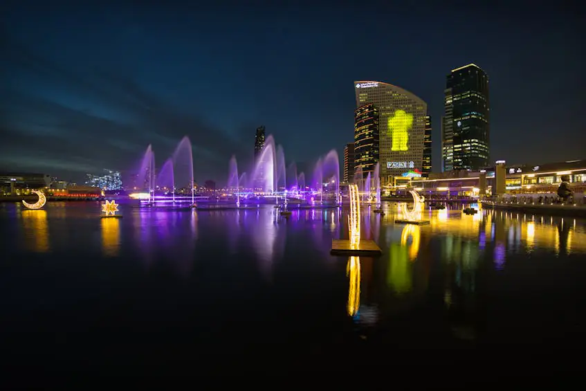 A stunning night view of a waterfront in Dubai, featuring illuminated fountains, modern high-rise buildings, and vibrant city lights. This image reflects the contrast in Ajman vs Dubai real estate investment, showcasing Dubai’s luxury developments and high-end property market.